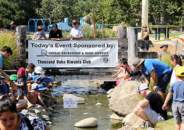 People gathered by a stream, signs for "Conejo Recreation & Park District" and "Thousand Oaks Kiwanis Club" in the background.