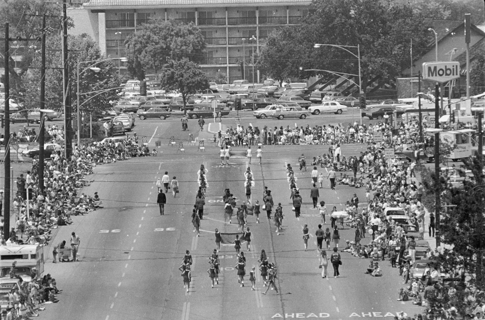Parade with people marching down a street, spectators on the sidelines, and vintage cars in the background.