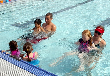 A swim class with two adults and several children in a pool, practicing swimming near the edge.