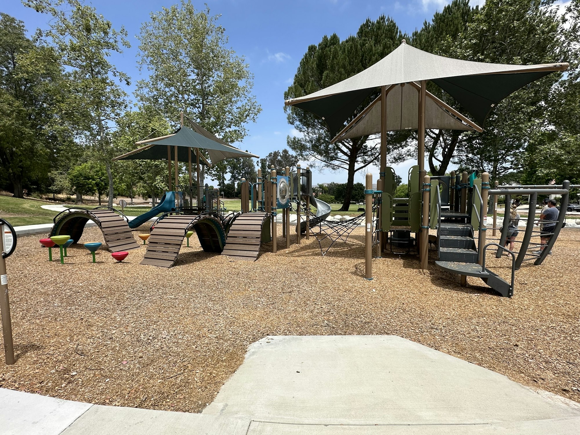 A playground featuring slides, climbing structures, and shaded areas, surrounded by trees and a gravel surface.