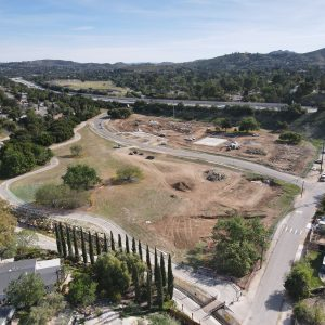 An aerial view of a construction site with roads, surrounding trees, and hills in the background.