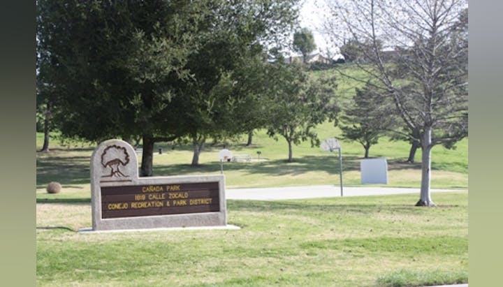 A park with a sign for "Cañada Park" and "Conejo Recreation & Park District," trees, grass, and a basketball court.