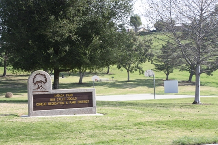 A park with a sign for "Cañada Park" and "Conejo Recreation & Park District," trees, grass, and a basketball court.