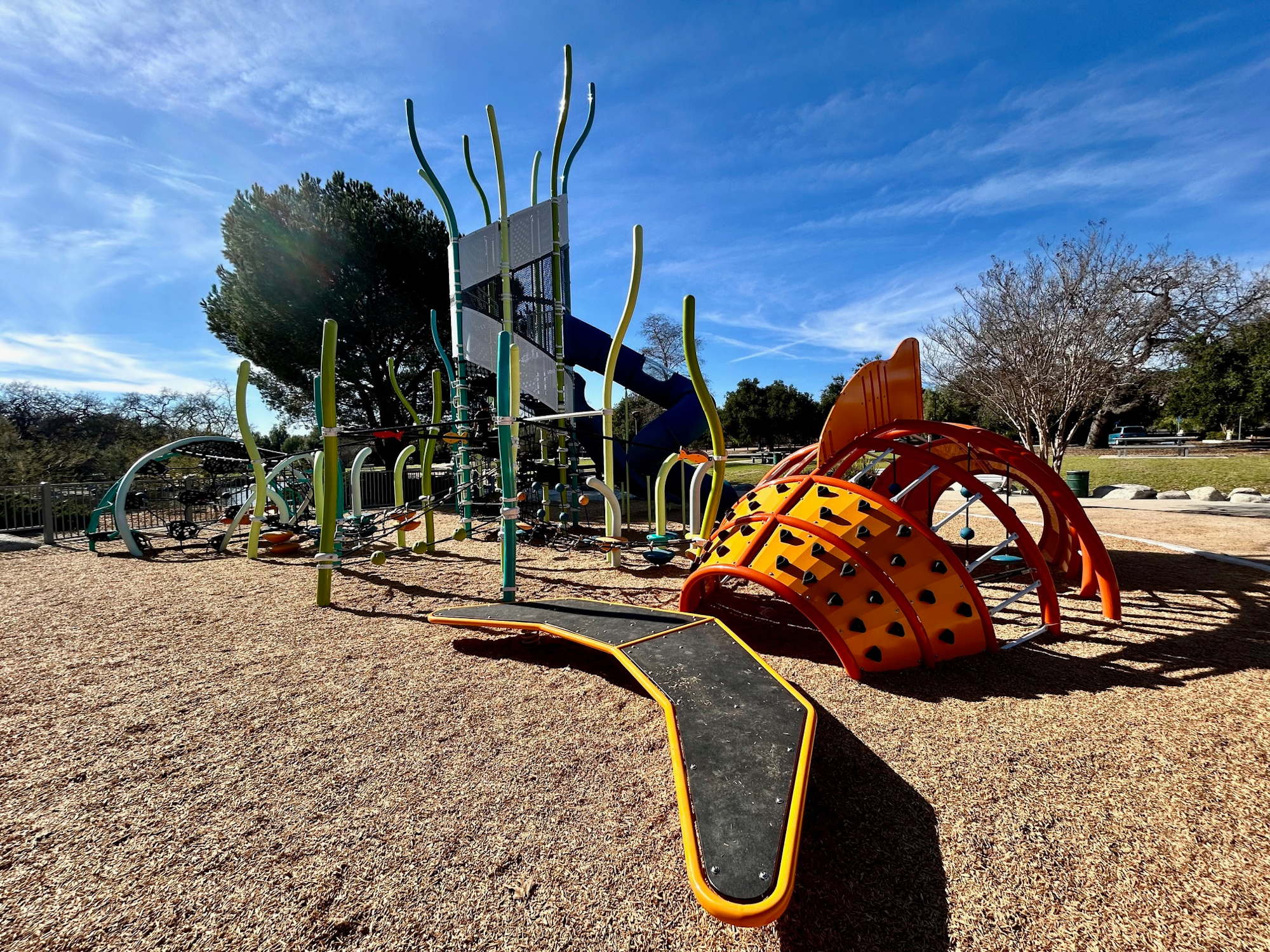 A modern playground with climbing structures, slides, and colorful equipment on a bark-covered surface under a clear blue sky.