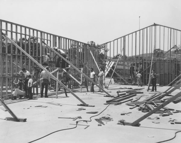 People constructing a wooden frame structure on a concrete foundation, with scattered tools and materials around.