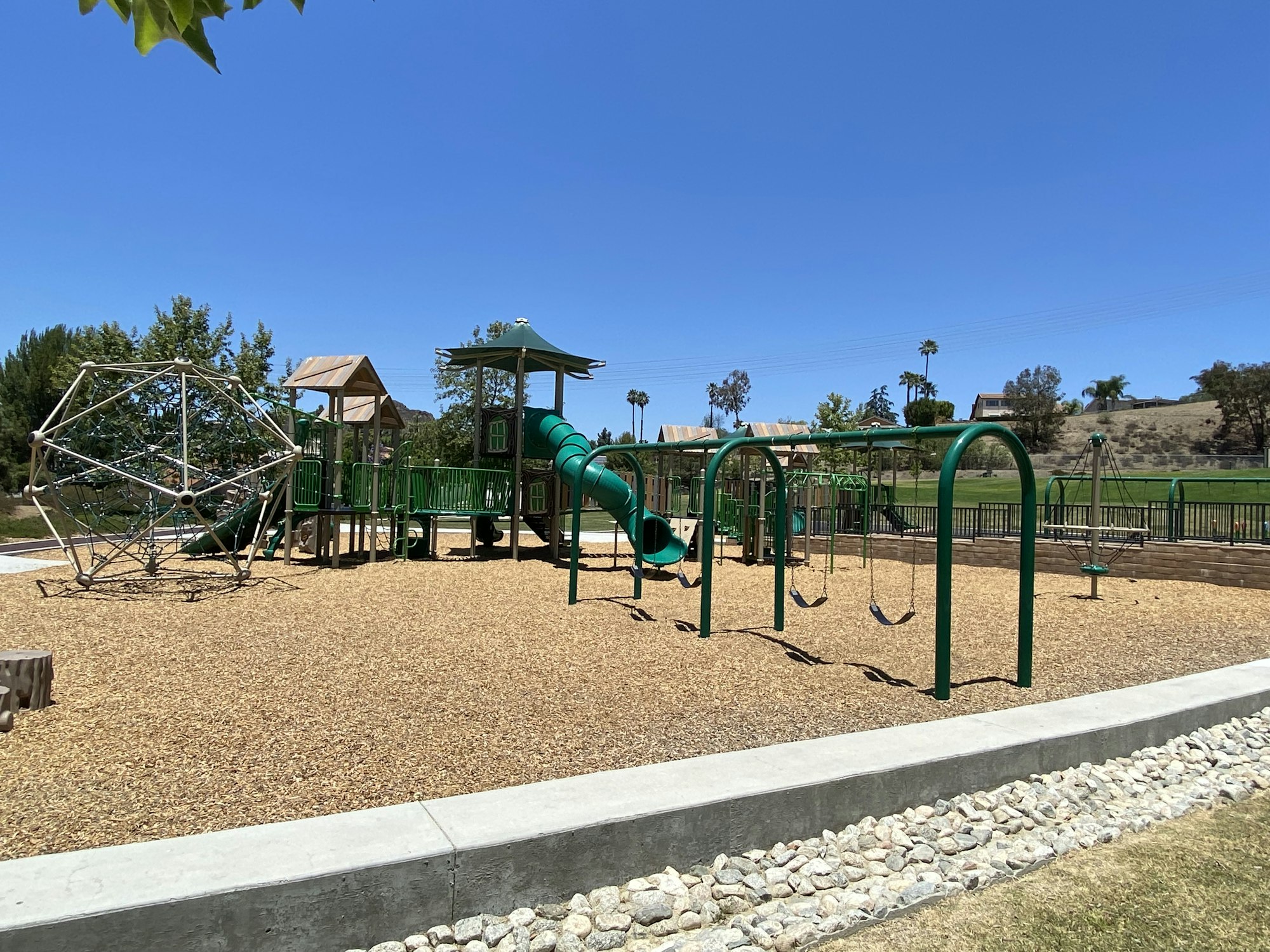 A playground with climbing structures, slides, swings, and a wood chip ground, surrounded by grassy areas and trees under a clear sky.