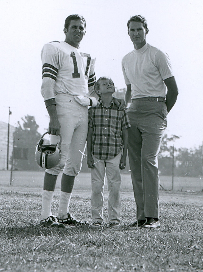 Three people standing together: a football player holding a helmet, a child in the center, and another man on the right.