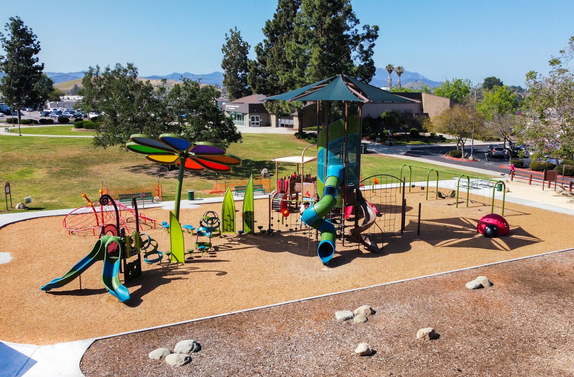 Colorful playground with slides, climbing structures, and a large flower-shaped shade. Trees and buildings are in the background.