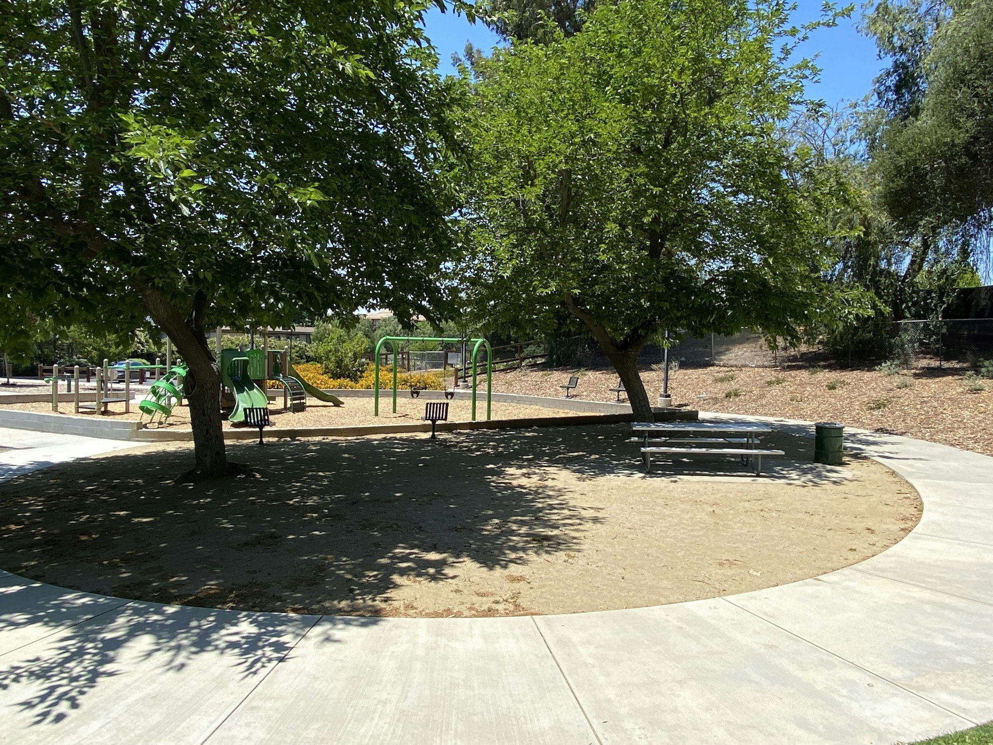 Playground with slides and swings, surrounded by trees and benches; concrete pathway in the foreground.