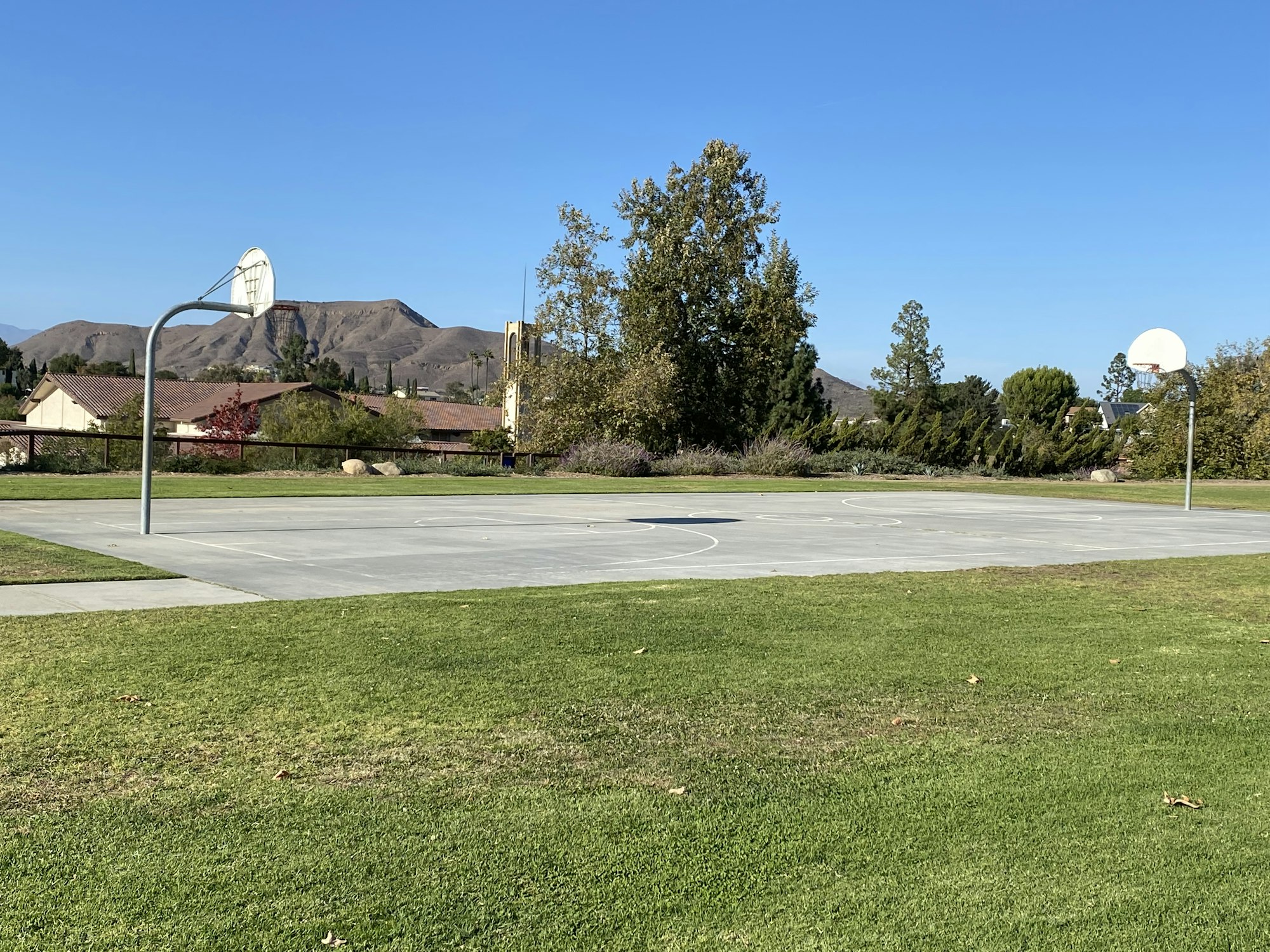 Outdoor basketball court surrounded by grass, trees, and hills in the background.