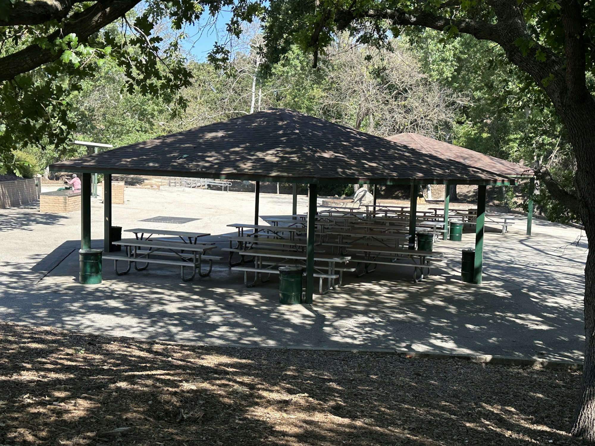 A park pavilion with picnic tables and trash cans, surrounded by trees and sunlight.