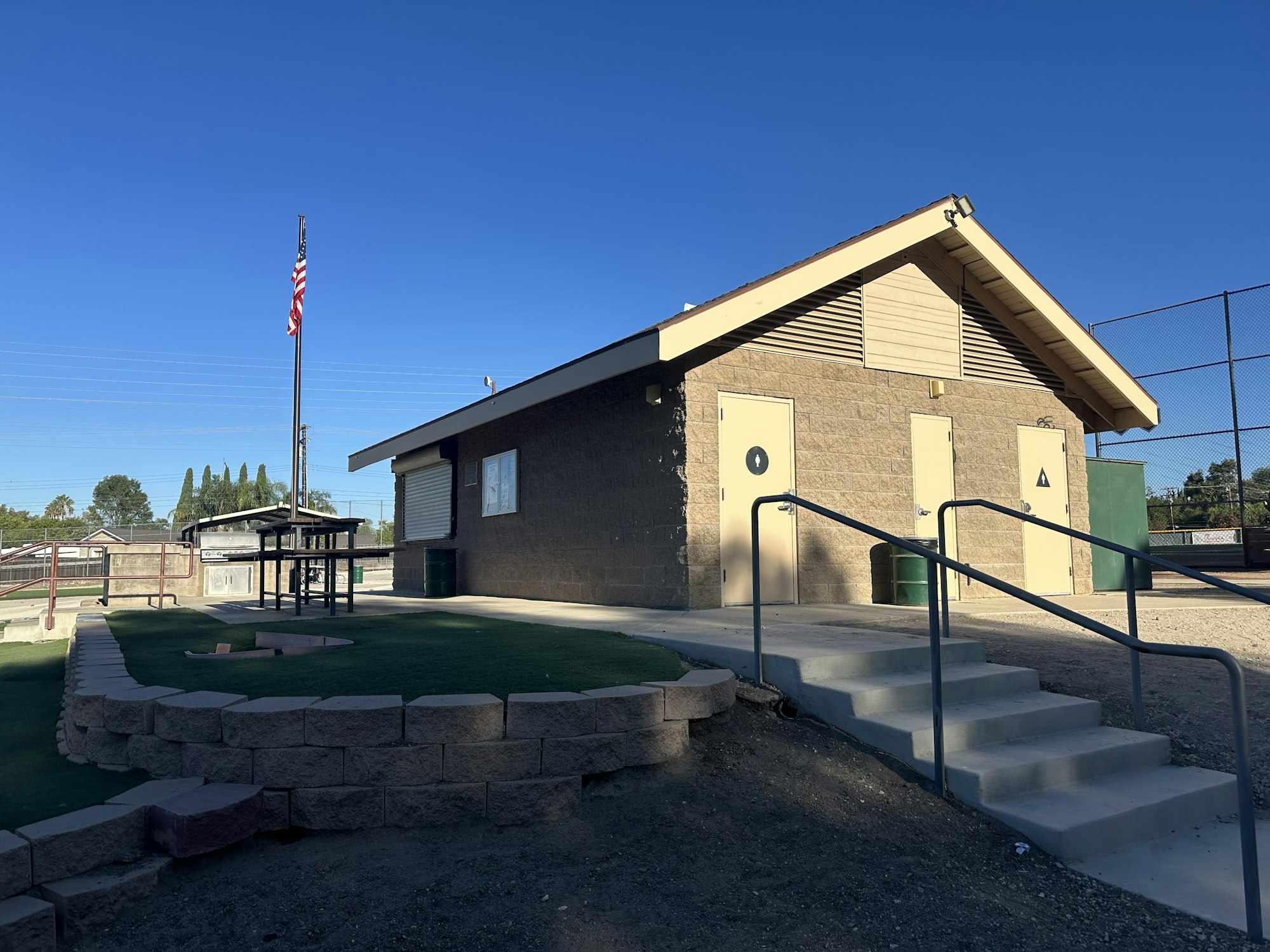 A small building with restrooms, a flagpole, and a picnic area, situated in a park setting under a clear blue sky.