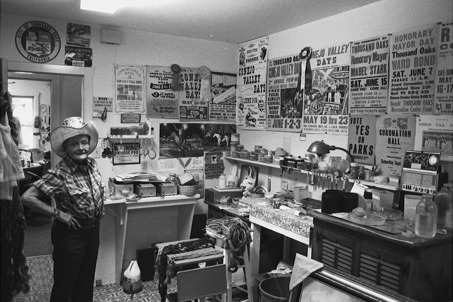 An elderly person in a cowboy hat stands in a cluttered room filled with posters and items related to Conejo Valley Days.