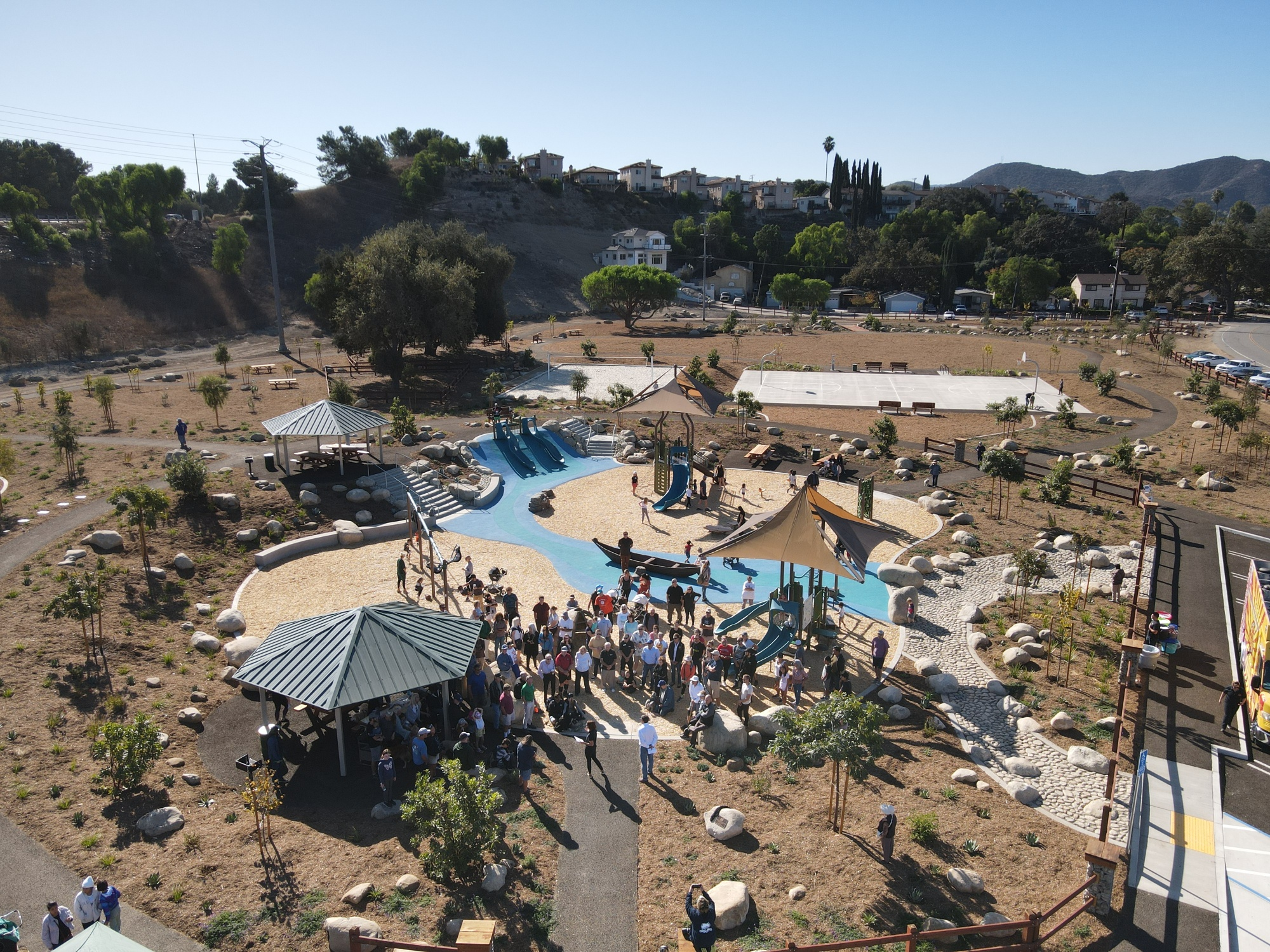 Aerial view of a playground with slides, shade structures, and people gathered, surrounded by grassy areas and trees.