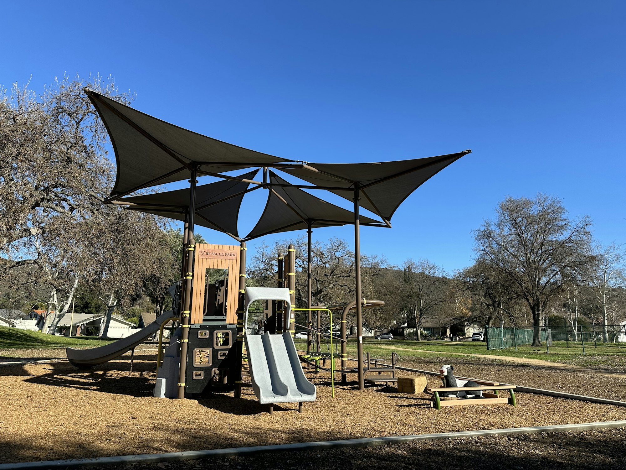 A playground with slides and climbing structures, covered by a sunshade, set in a park with trees and clear blue sky.