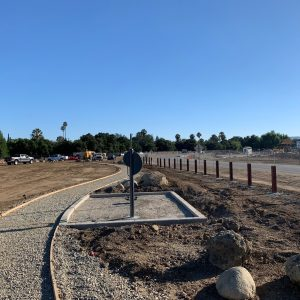 Under-construction site with a gravel path, utility posts, and scattered vehicles. Trees and a clear sky in the background.