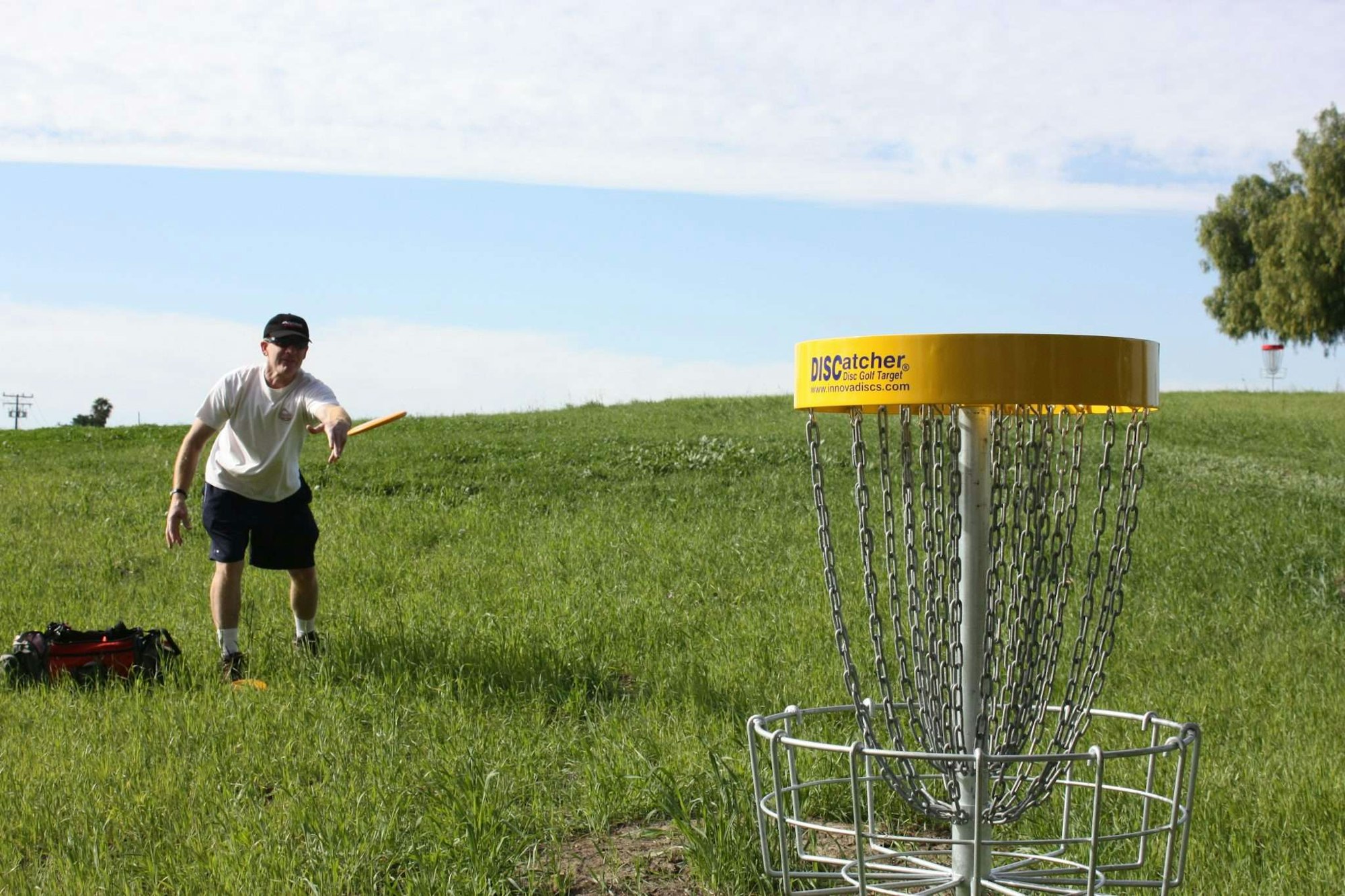 A person is playing disc golf, aiming a disc at a disc golf basket in a grassy field.
