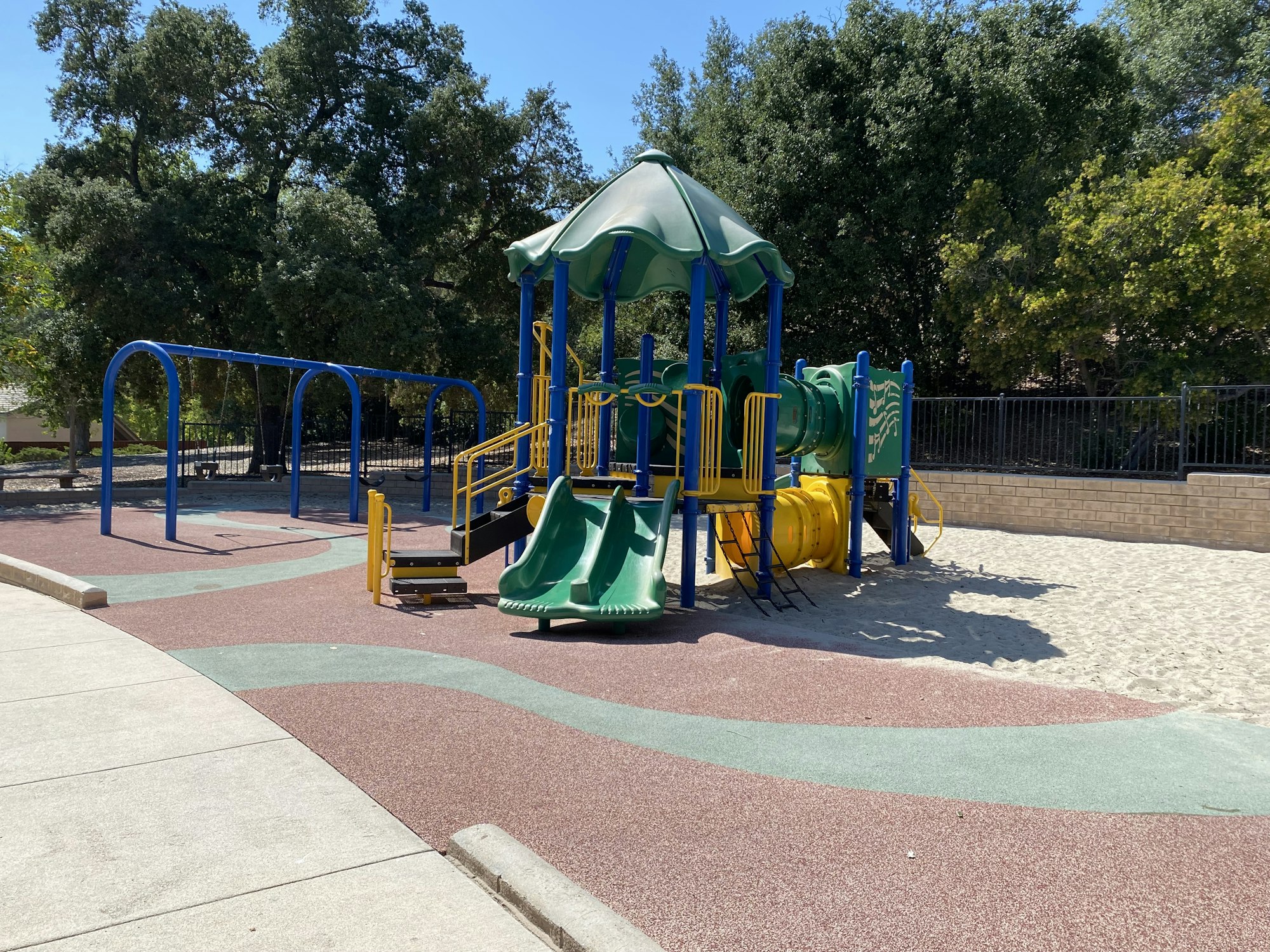 Playground with slides, swings, and climbing structures in a park setting with trees and a sandy area.