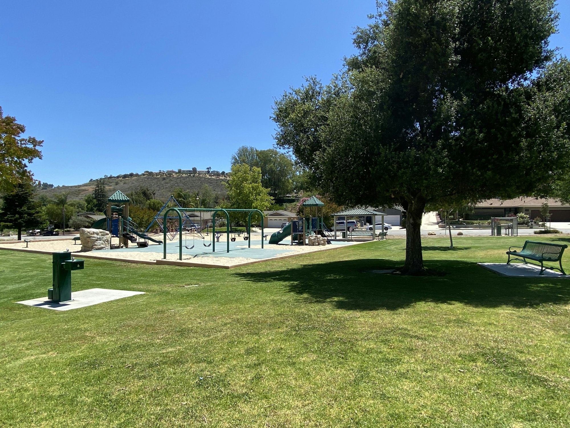 A sunny park with a playground, swings, a tree, benches, and grassy area.