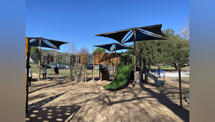 Playground with slides, climbing structures, and shade canopies. Children and adults present, clear blue sky.