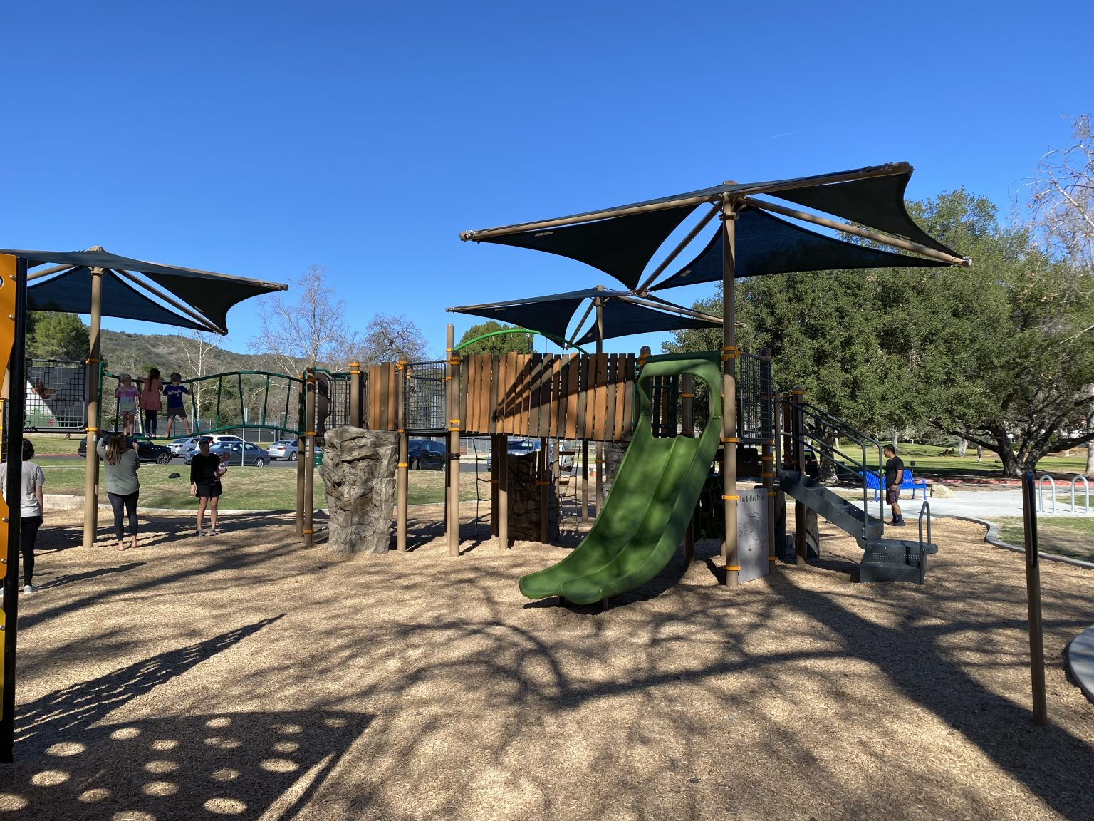 Playground with slides, climbing structures, and shade canopies. Children and adults present, clear blue sky.