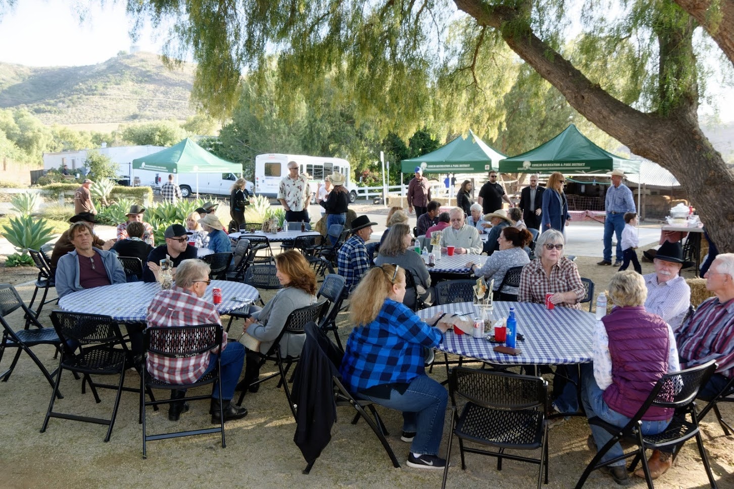 Outdoor gathering with people sitting at tables, green tents in the background, and trees providing shade.