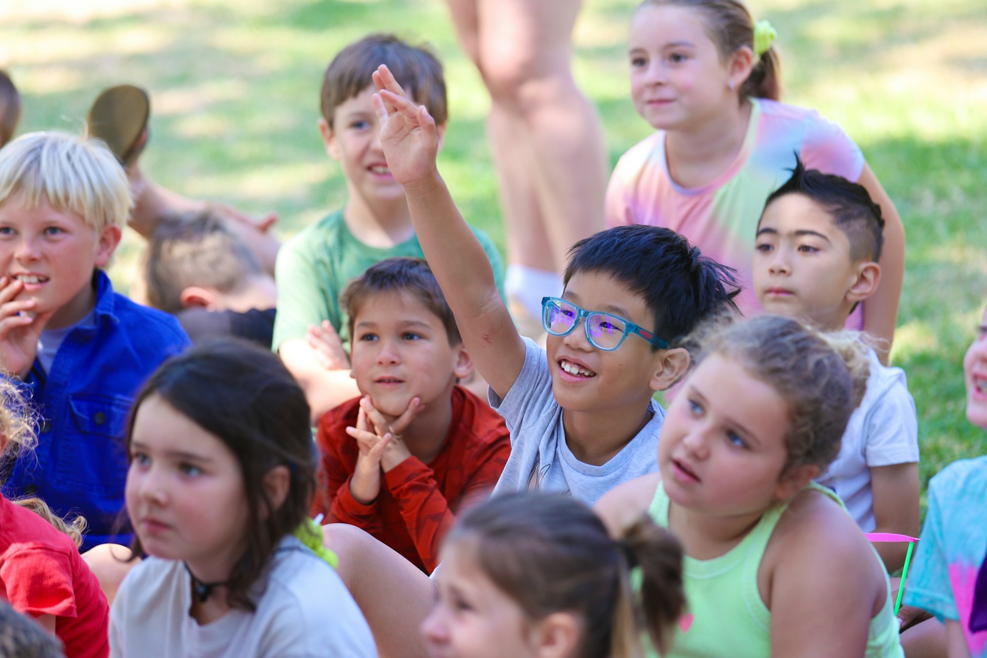A group of children sits outside. A child in glasses raises his hand eagerly.