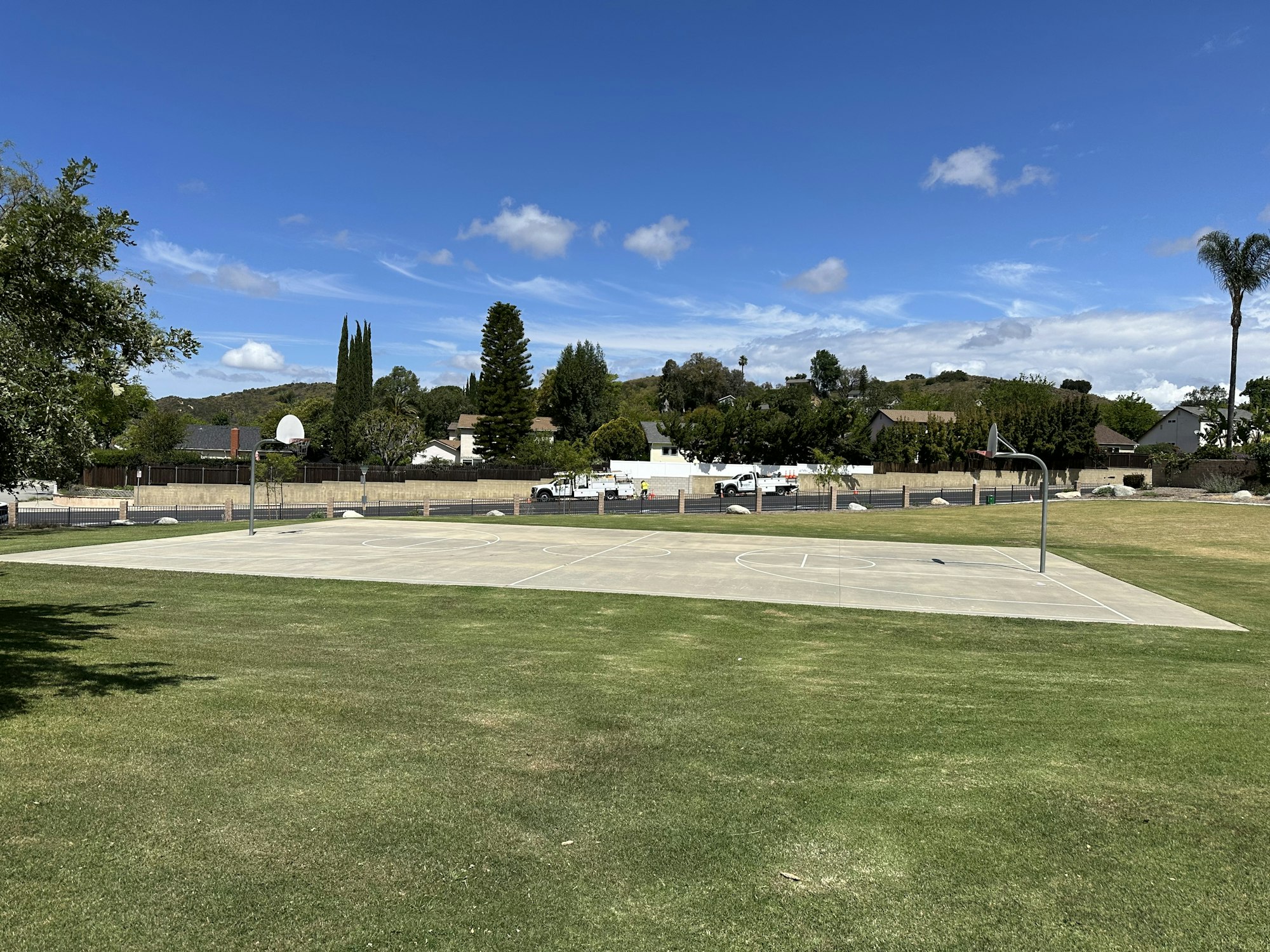 A spacious outdoor basketball court surrounded by grass, trees, and a clear blue sky, with houses in the background.
