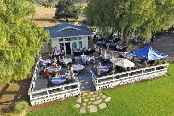 People are gathered at outdoor tables on a patio surrounded by trees, next to a house-like building.
