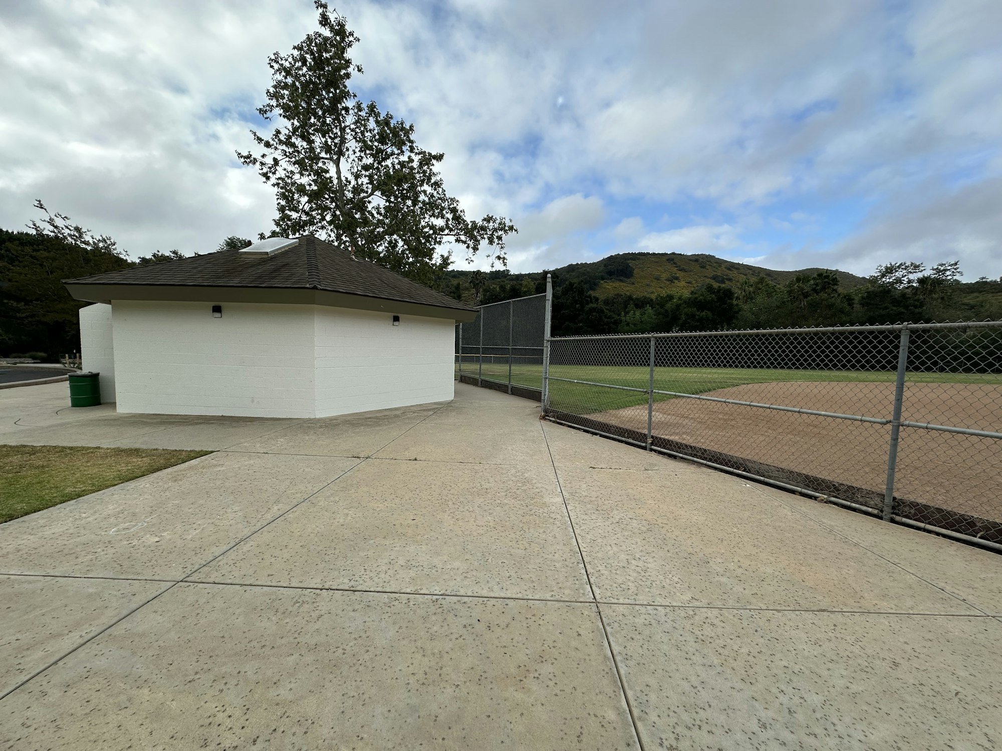 A white building near a baseball field, with a concrete path and a scenic hill in the background under a cloudy sky.