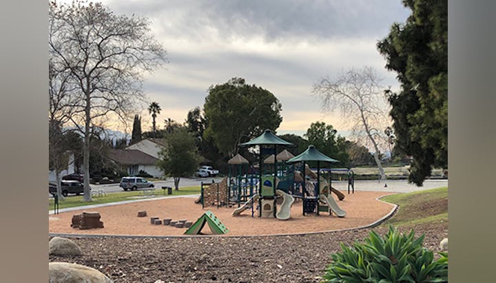 Playground with slides and climbing structures, surrounded by trees and houses under a cloudy sky.
