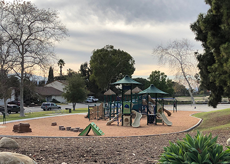 Playground with slides and climbing structures, surrounded by trees and houses under a cloudy sky.