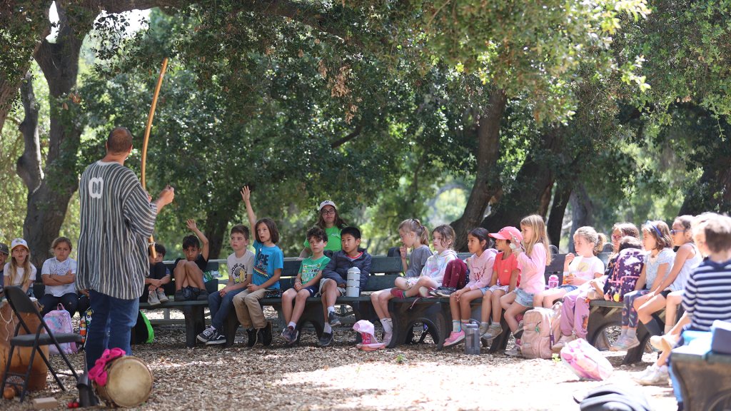 A person teaching a group of children outdoors. The kids sit on benches under trees, some with backpacks, attentive to the lesson.
