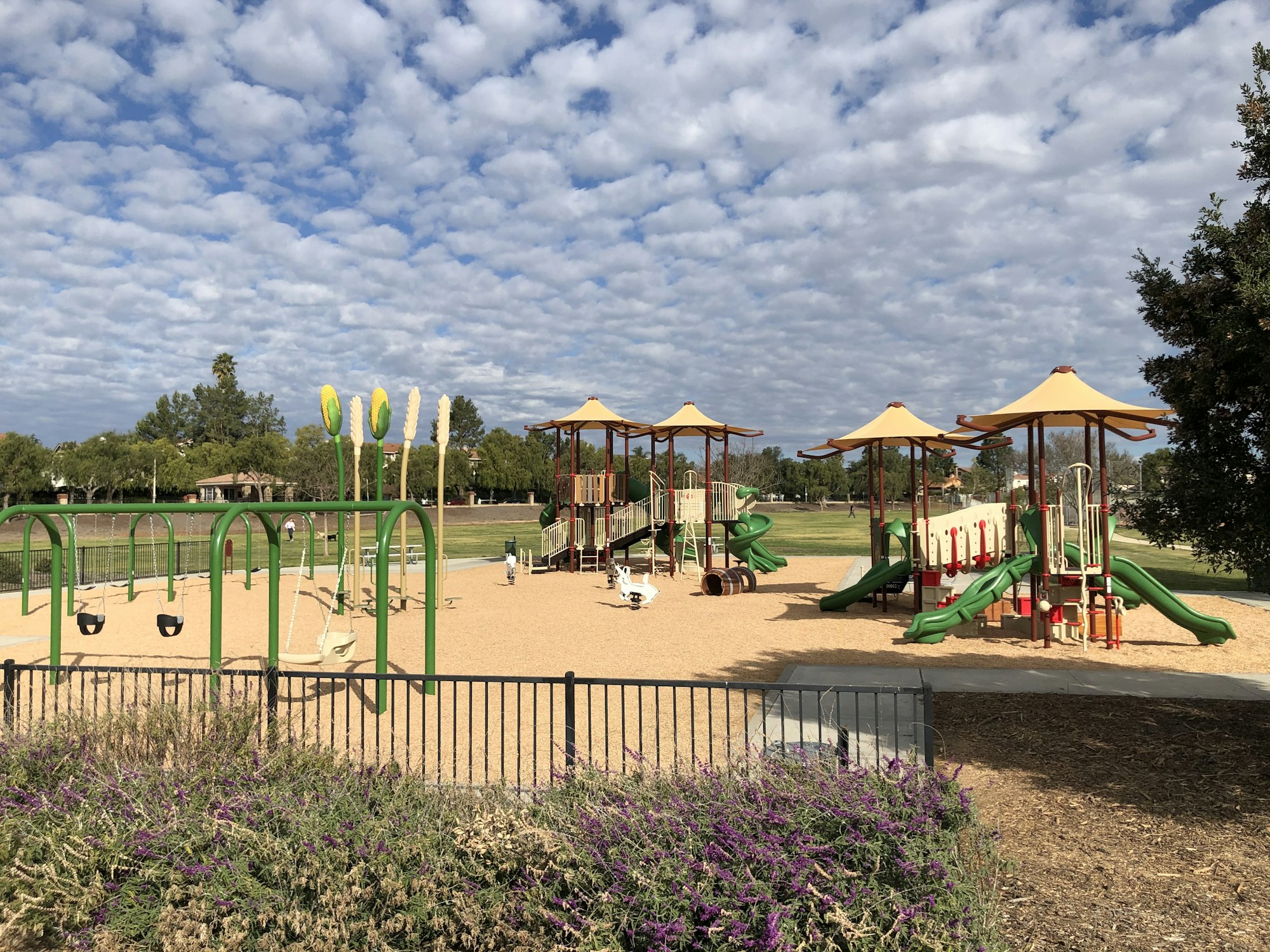 Playground with slides, swings, climbing structures, surrounded by a fence and greenery under a partly cloudy sky.