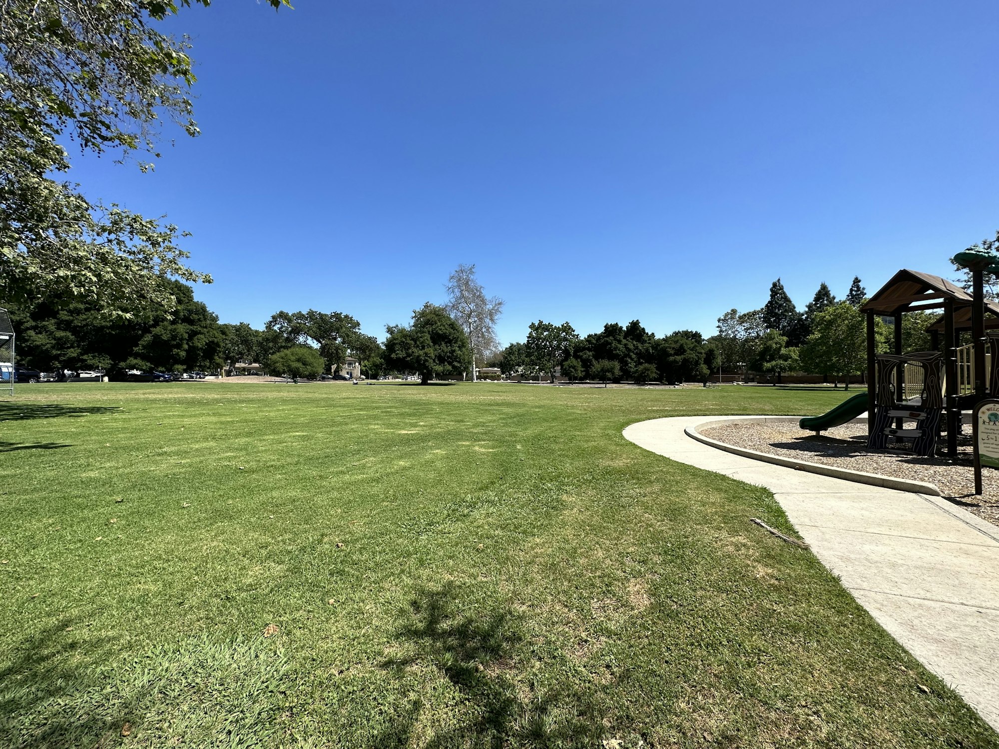 A park with a grassy field, trees, and a playground structure under a clear blue sky.