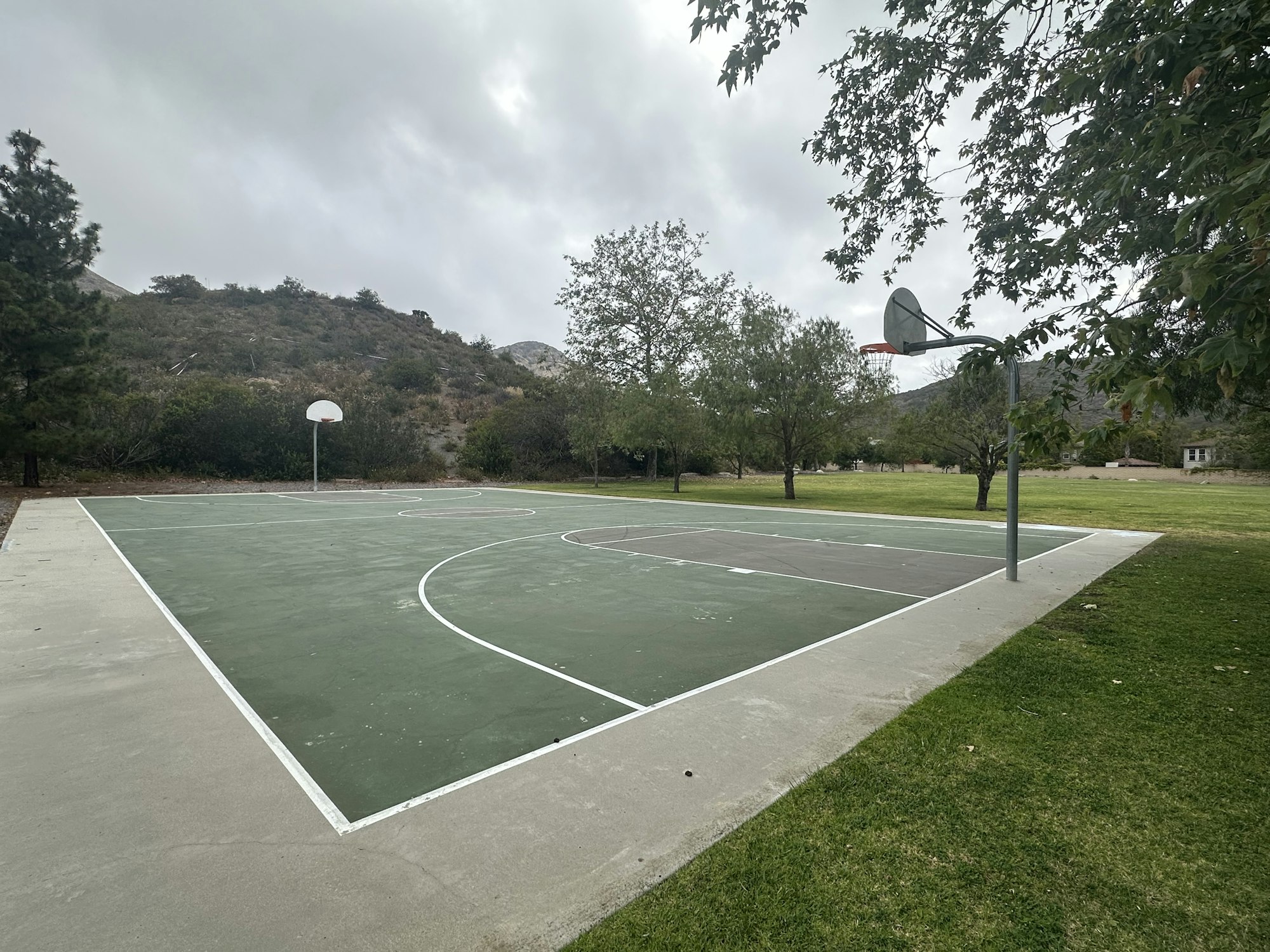 An empty outdoor basketball court surrounded by trees and grass, with mountains in the background under a cloudy sky.