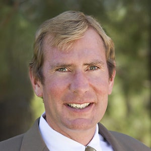 A man in a suit smiling against a blurred, natural background.
