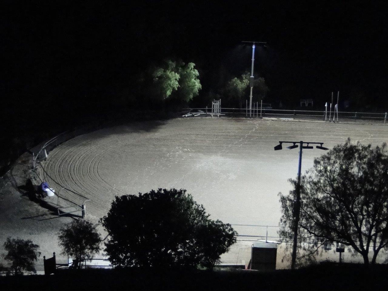 An illuminated outdoor arena at night, surrounded by trees, with visible tracks on the ground and a few lighting poles.