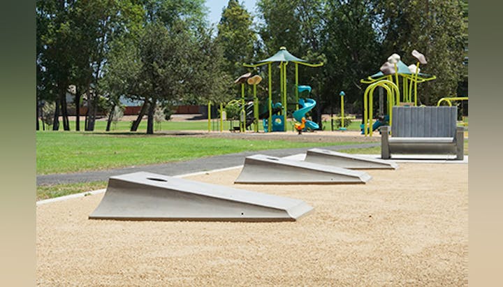 A skate park area with ramps and a bench, near a playground with climbing structures and slides, surrounded by trees.