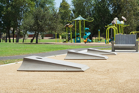 A skate park area with ramps and a bench, near a playground with climbing structures and slides, surrounded by trees.