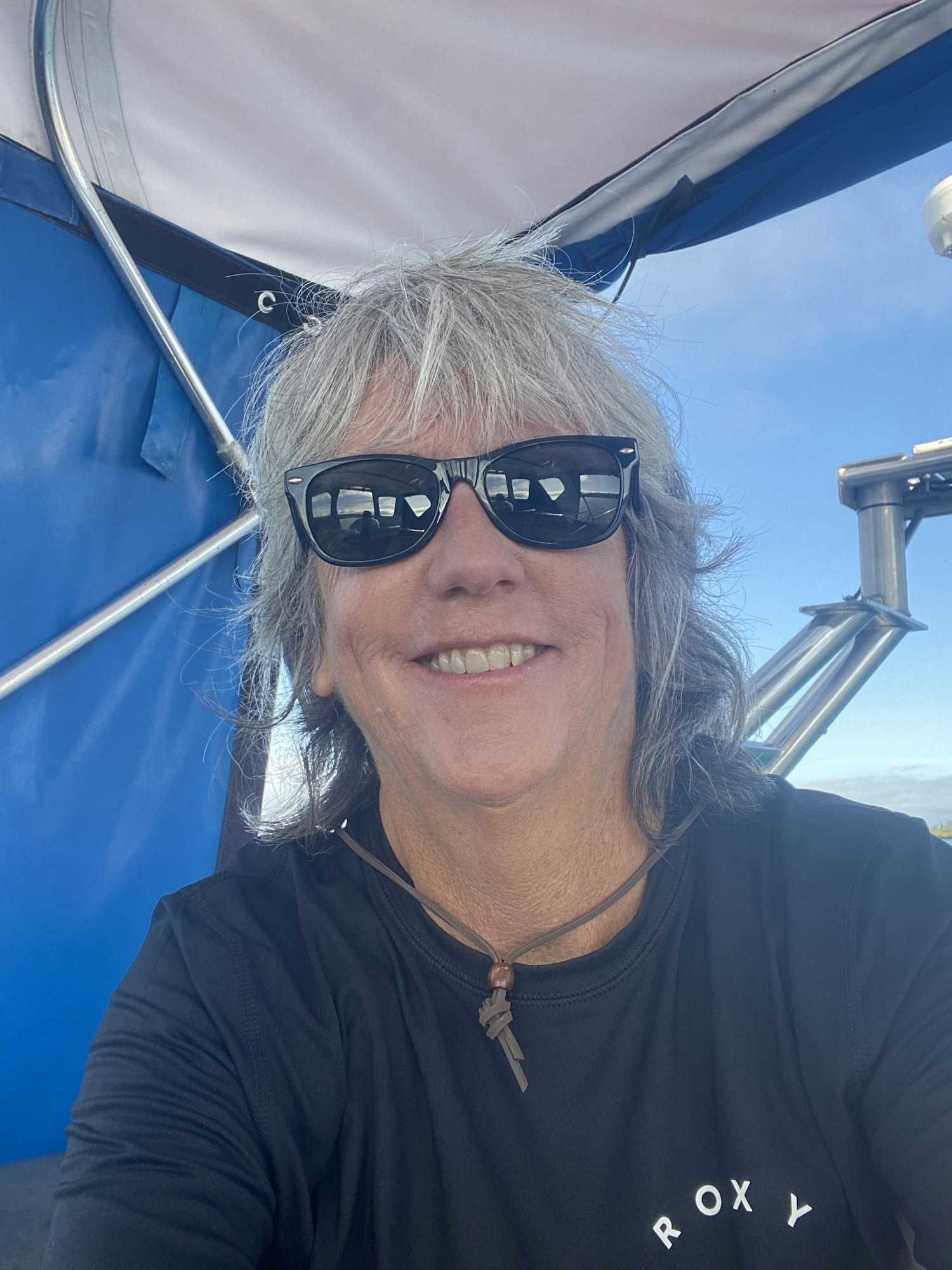 A person with grey hair, wearing sunglasses and a black shirt, smiles in a boat setting under a blue sky.