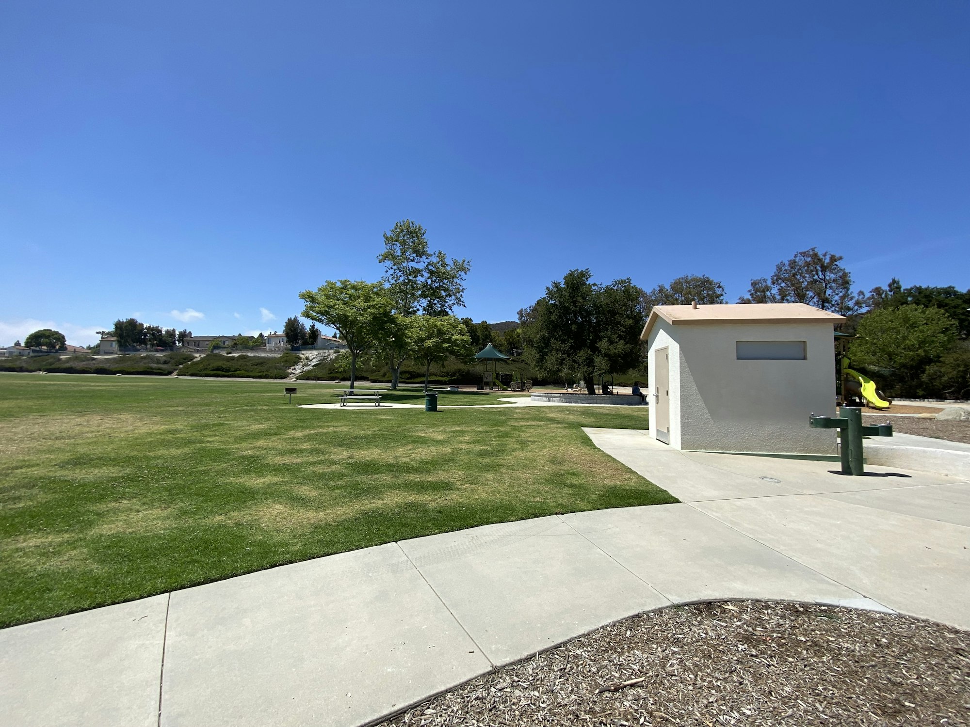 A sunny park with a grassy field, trees, a small building, pathways, and playground equipment.