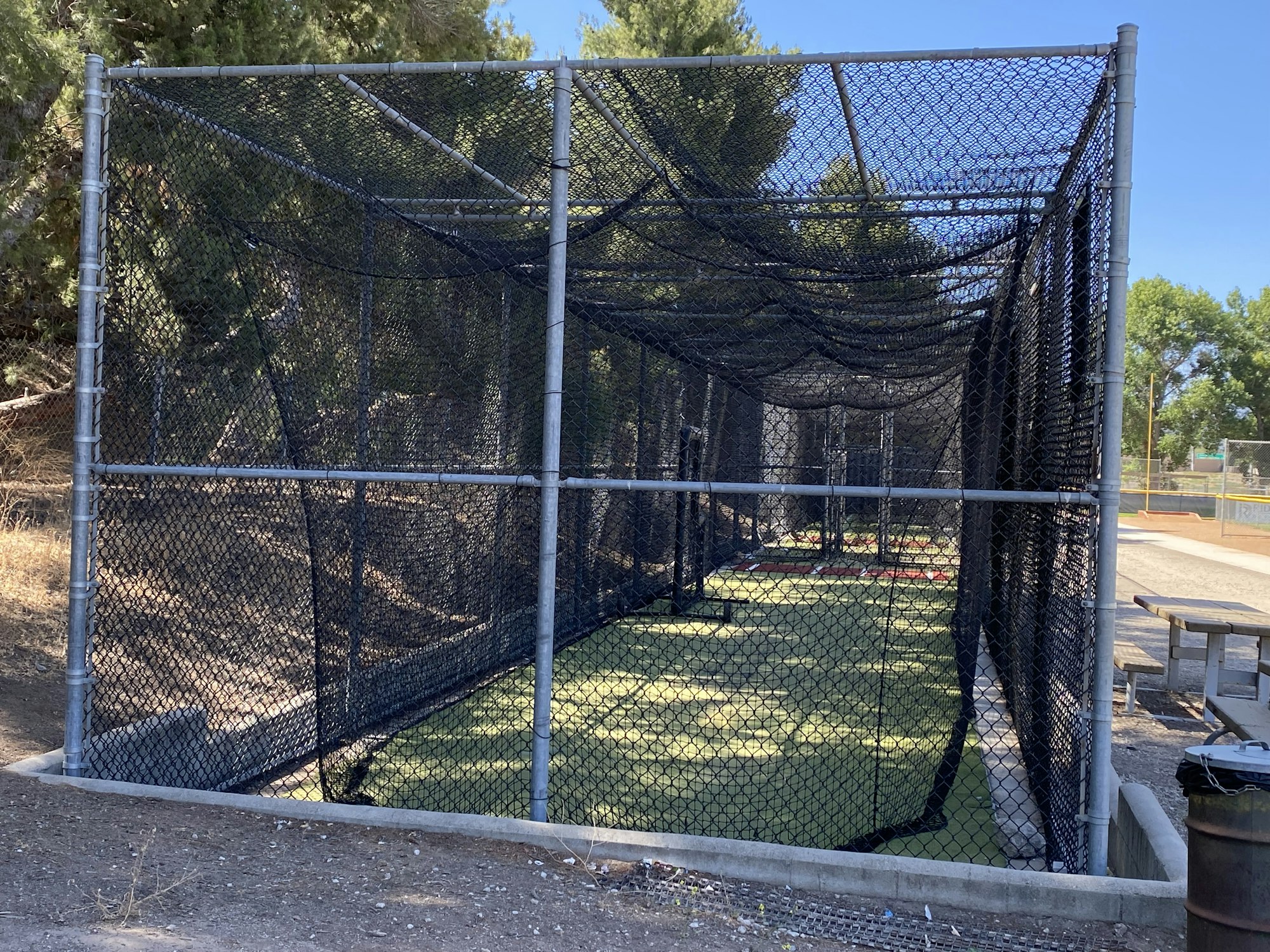 A baseball batting cage surrounded by trees with a picnic table and trash can nearby.