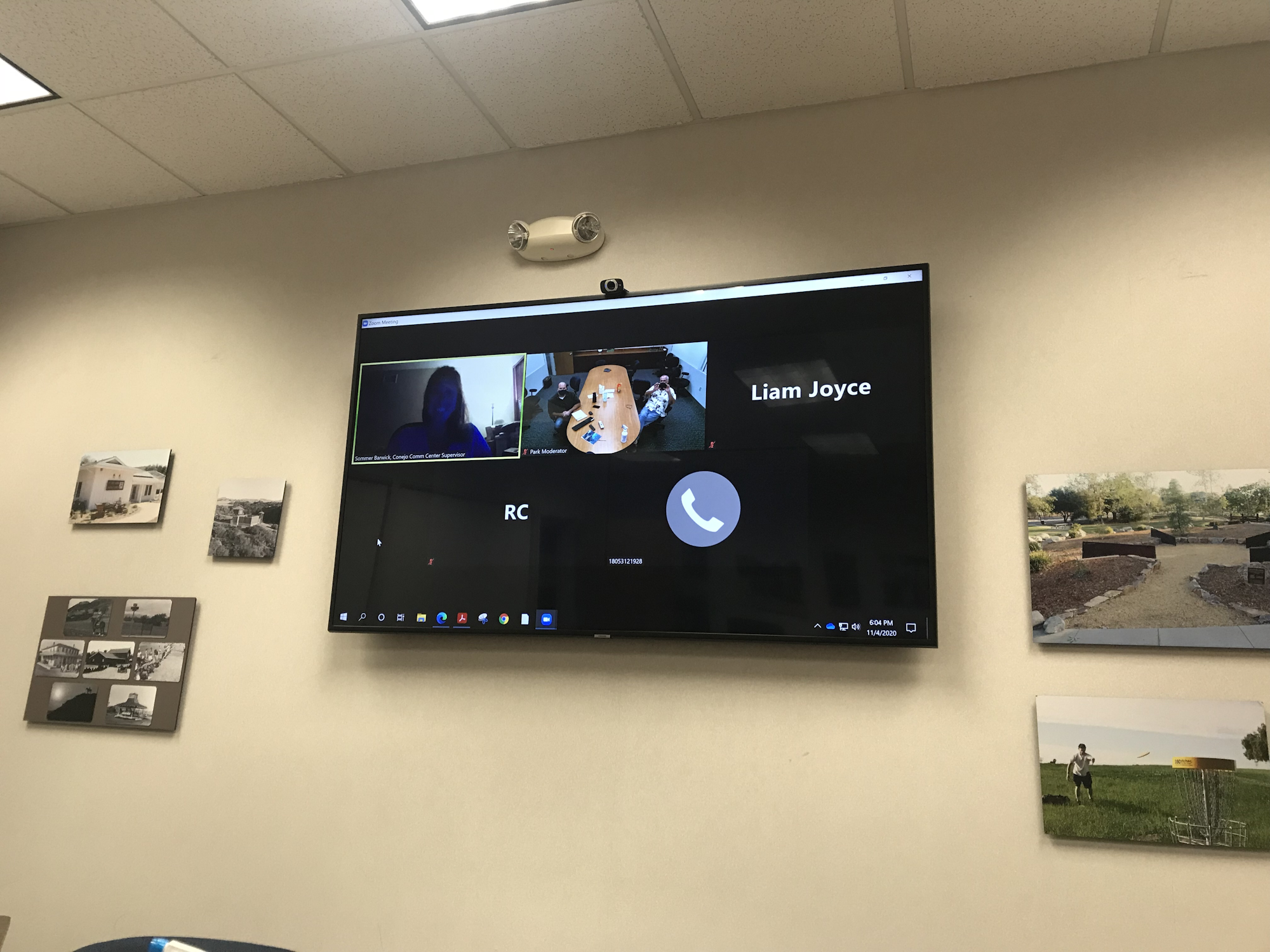 Video conference on a wall-mounted screen in an office with framed photos.