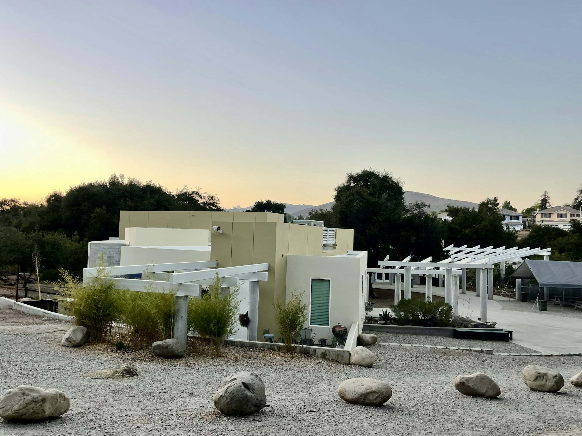 Modern house with flat roofs and white pergolas, surrounded by gravel and rocks, with trees and a sunset in the background.
