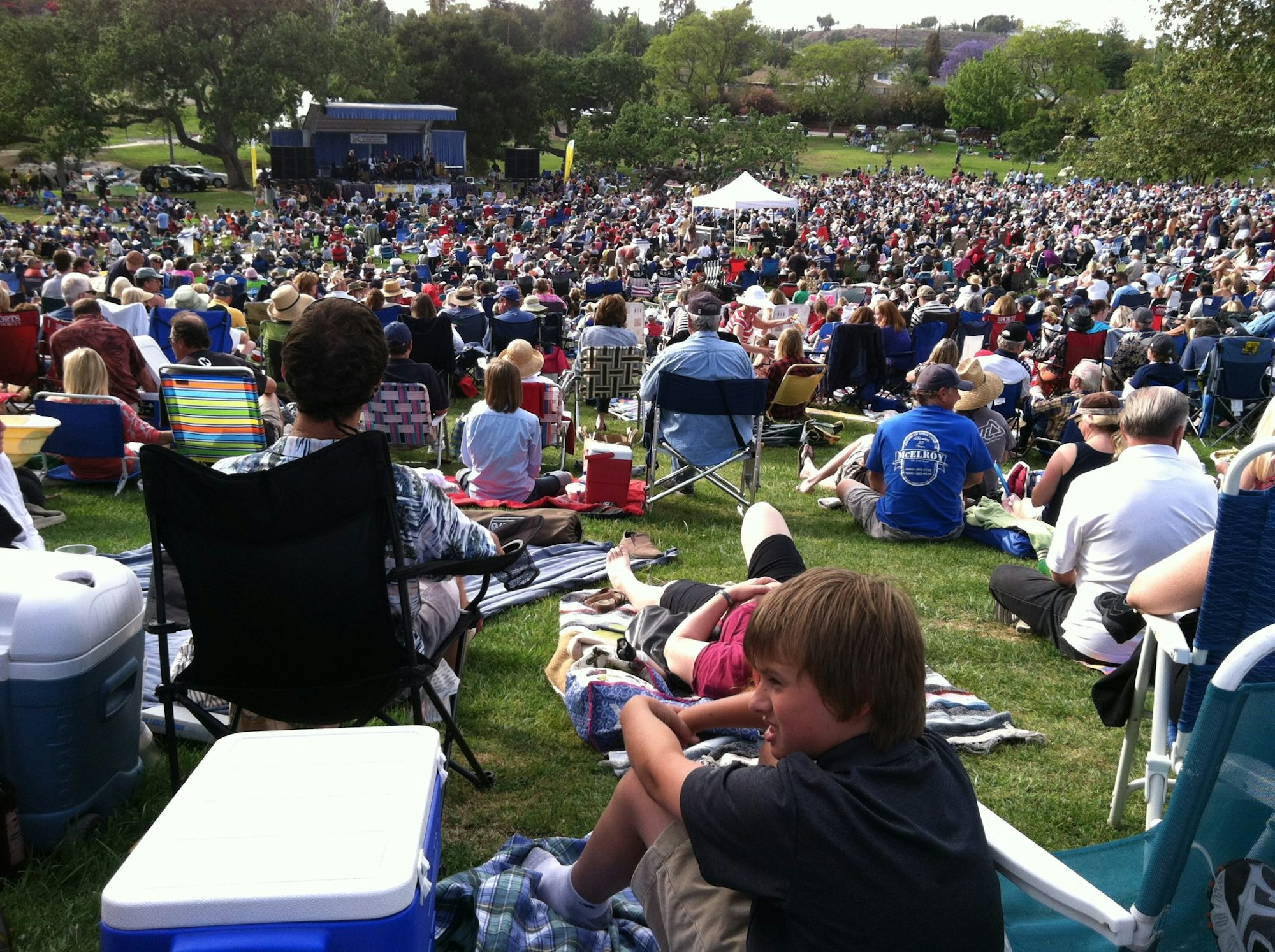 Crowd gathered on grass with blankets and chairs at an outdoor concert or event, stage in the distance.