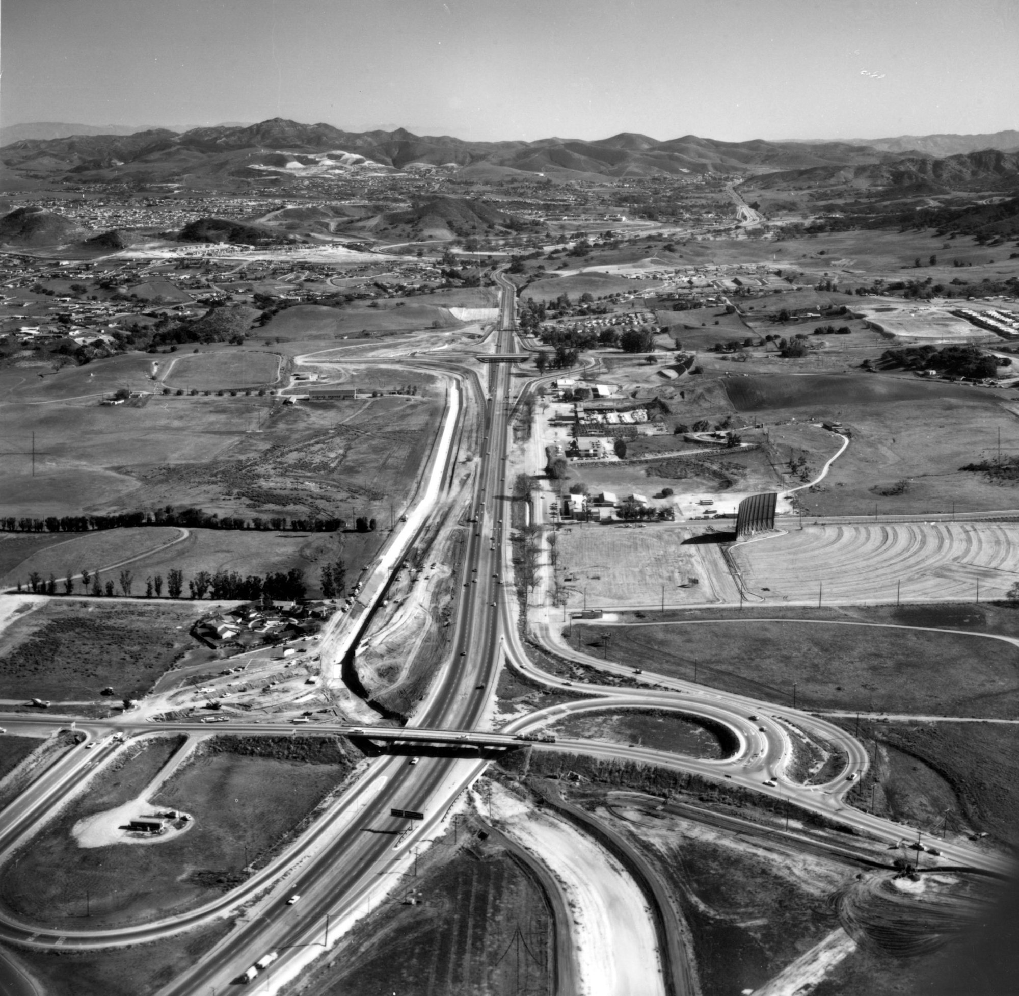 Aerial view of a highway interchange, open fields, scattered buildings, and distant hills under a clear sky.