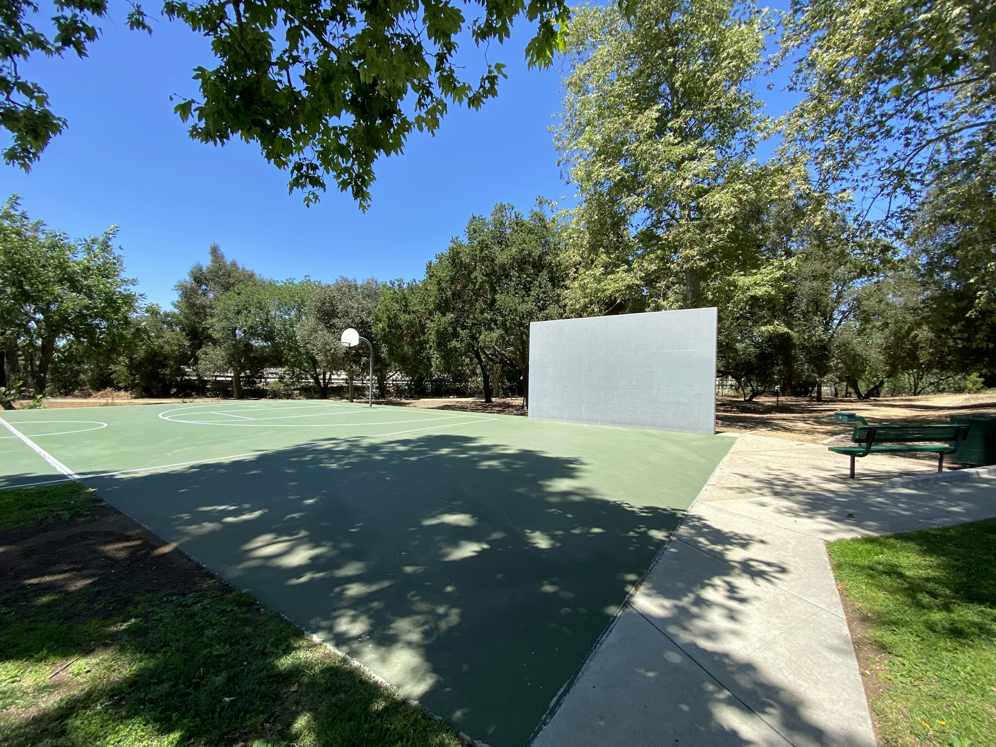 Outdoor basketball court with a backboard and wall, surrounded by trees on a sunny day.