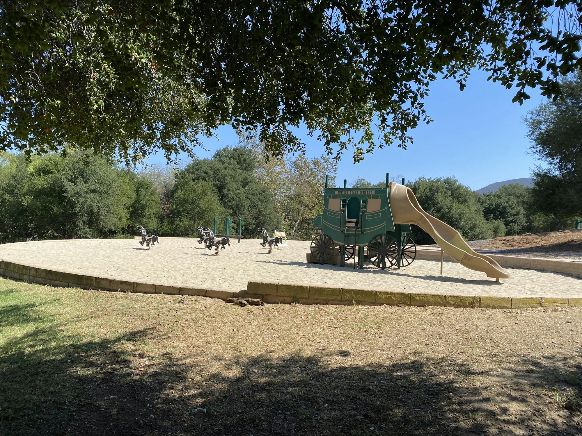 A playground with a stagecoach-themed slide and bouncy horses on a sandy area surrounded by trees.