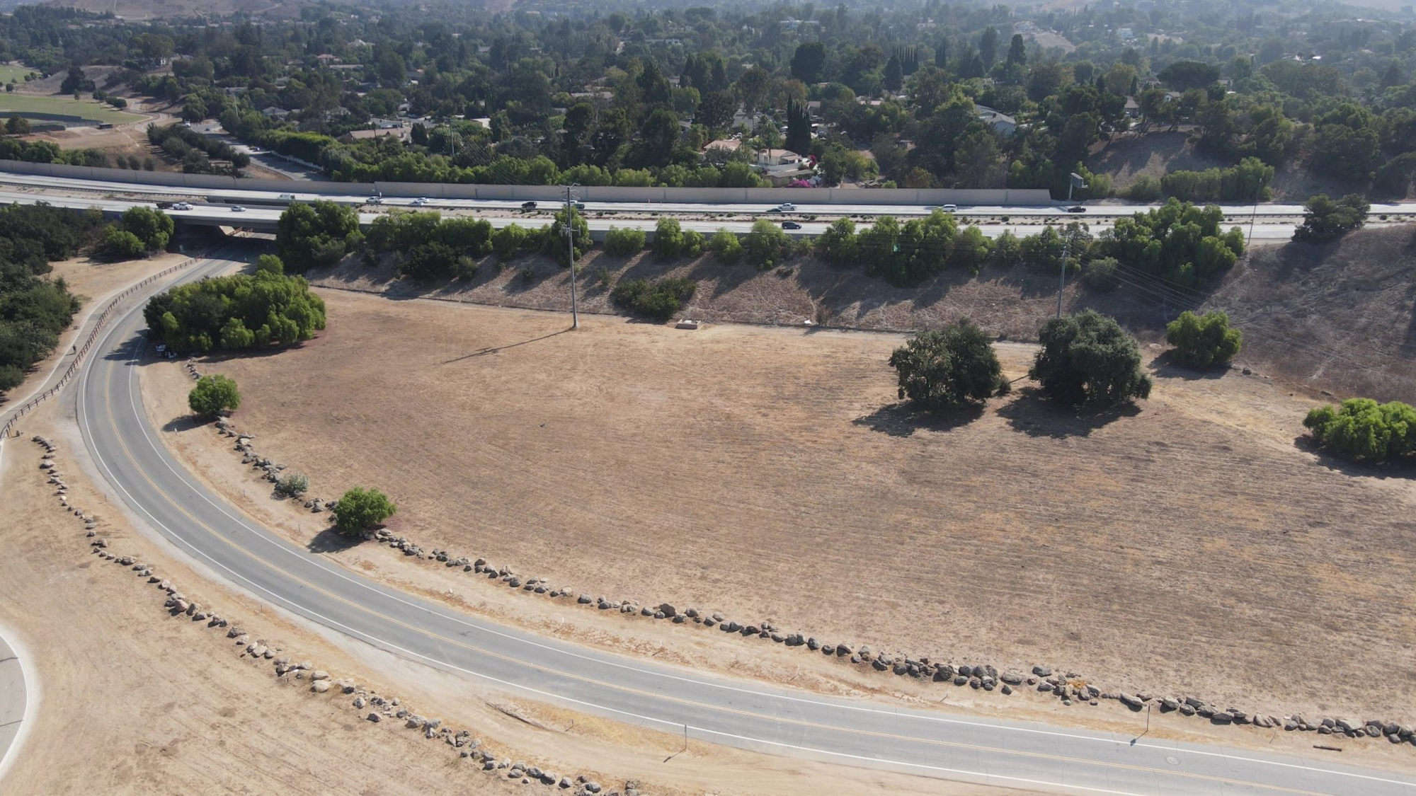 Curved road with rocks, dry grass field, trees, and highway with vehicles in the background.